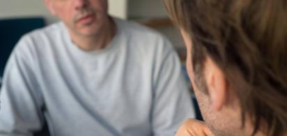 Zwei m&auml;nnlich gelesene Personen sitzen sich in einem ernsten Gespr&auml;ch gegen&uuml;ber.